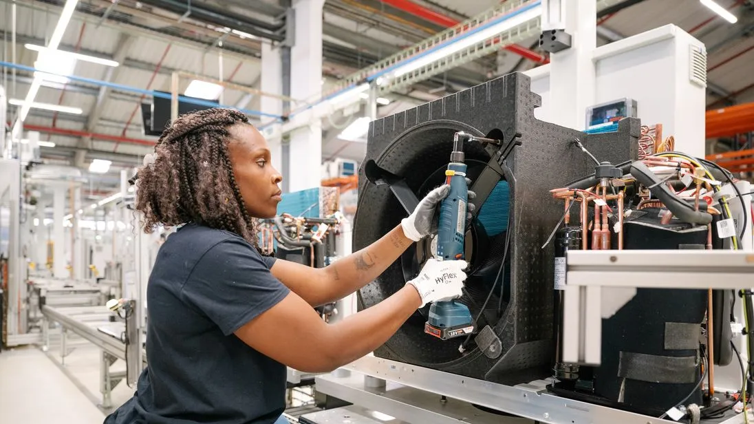 Eine Person montiert ein Bauteil an einer Wärmepumpe in einer Fabrikhalle. 